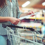 Woman with shopping cart in aisle checks and examines a sales receipt after purchasing food in a grocery store. Customer purchase products at supermarket.