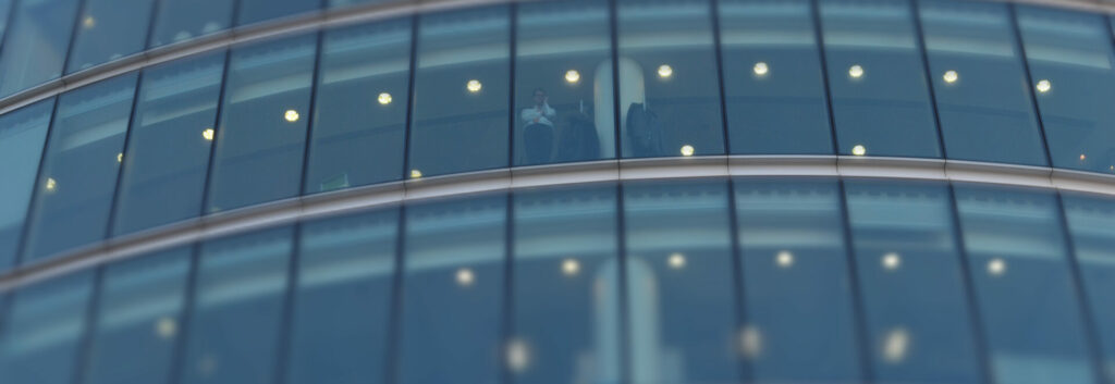 Photo of a businessman looking out of the window of a multistory office building, taken from outside the building.