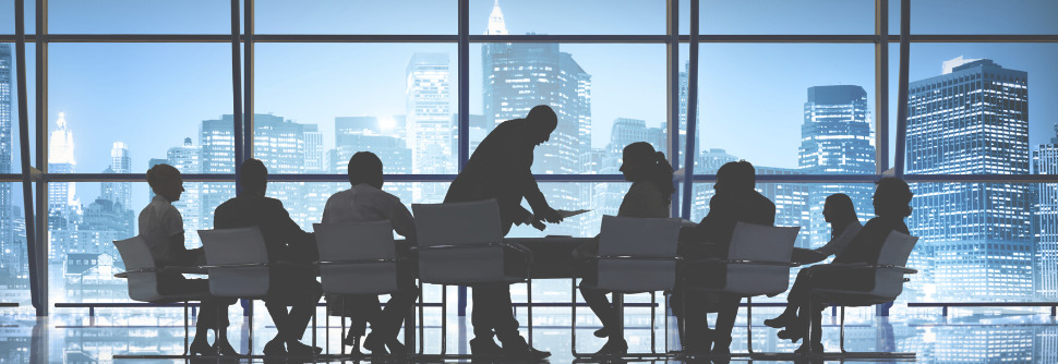 Silhouettes of business people meeting at a long table with a city skyline visible through windows behind them.