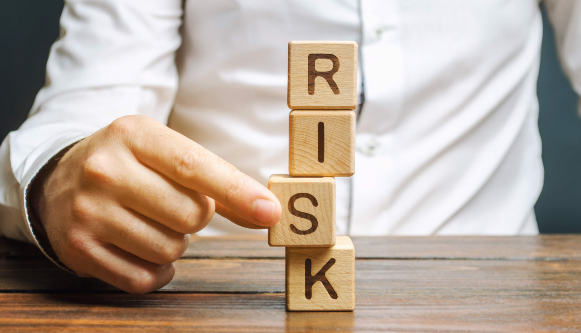 Hand holding a wooden block at the bottom of a stack. The blocks spell out 'RISK'